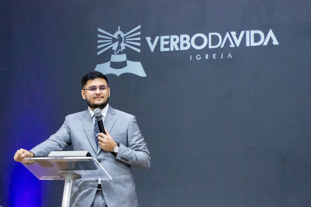 Daniel smiling at the pulpit in a grey suit, holding a microphone