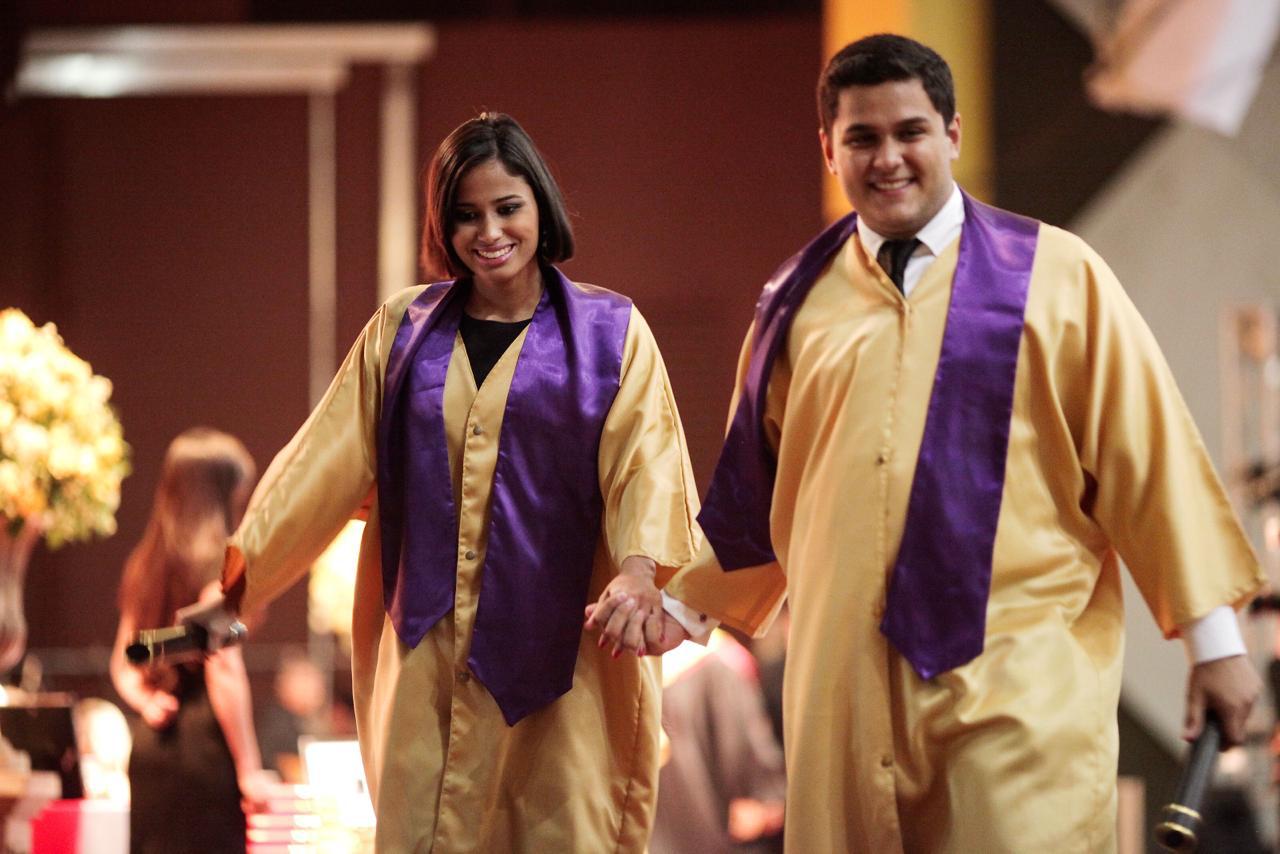 Daniel and Carla walking hand in hand in gold and purple gowns, each holding a diploma